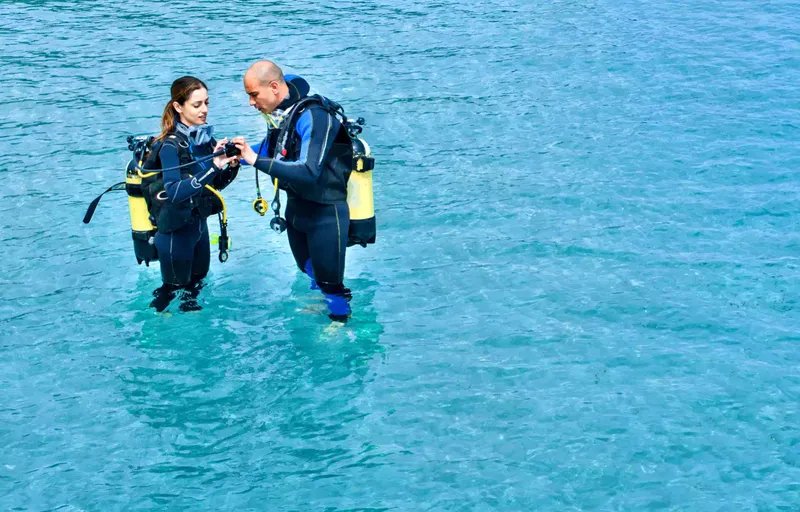 Un hombre y una mujer con equipo de buceo se encuentran en el agua hasta las rodillas e inspeccionan el rebreather de la mujer
