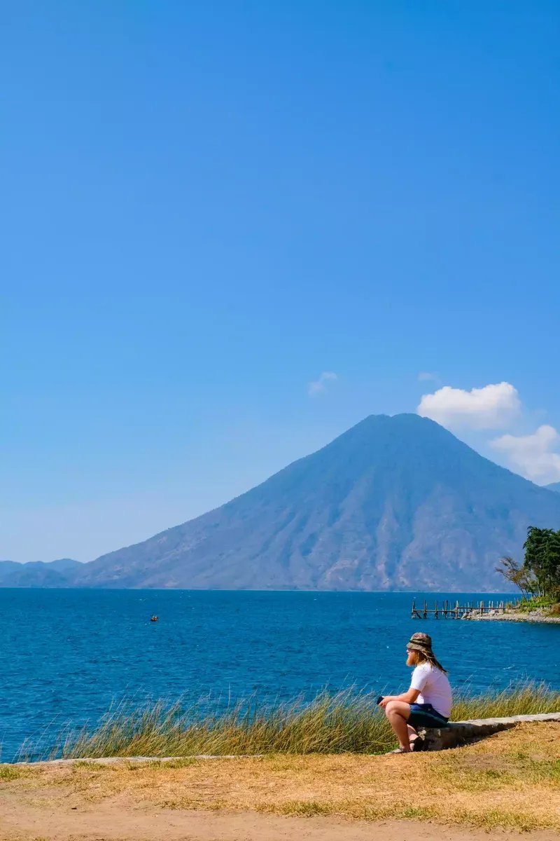Una persona sentada en la orilla del lago Atitlán con un volcán de fondo